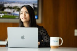 A young woman works on a laptop at a desk, with an LMU mug beside her and campus imagery visible on a screen behind her.