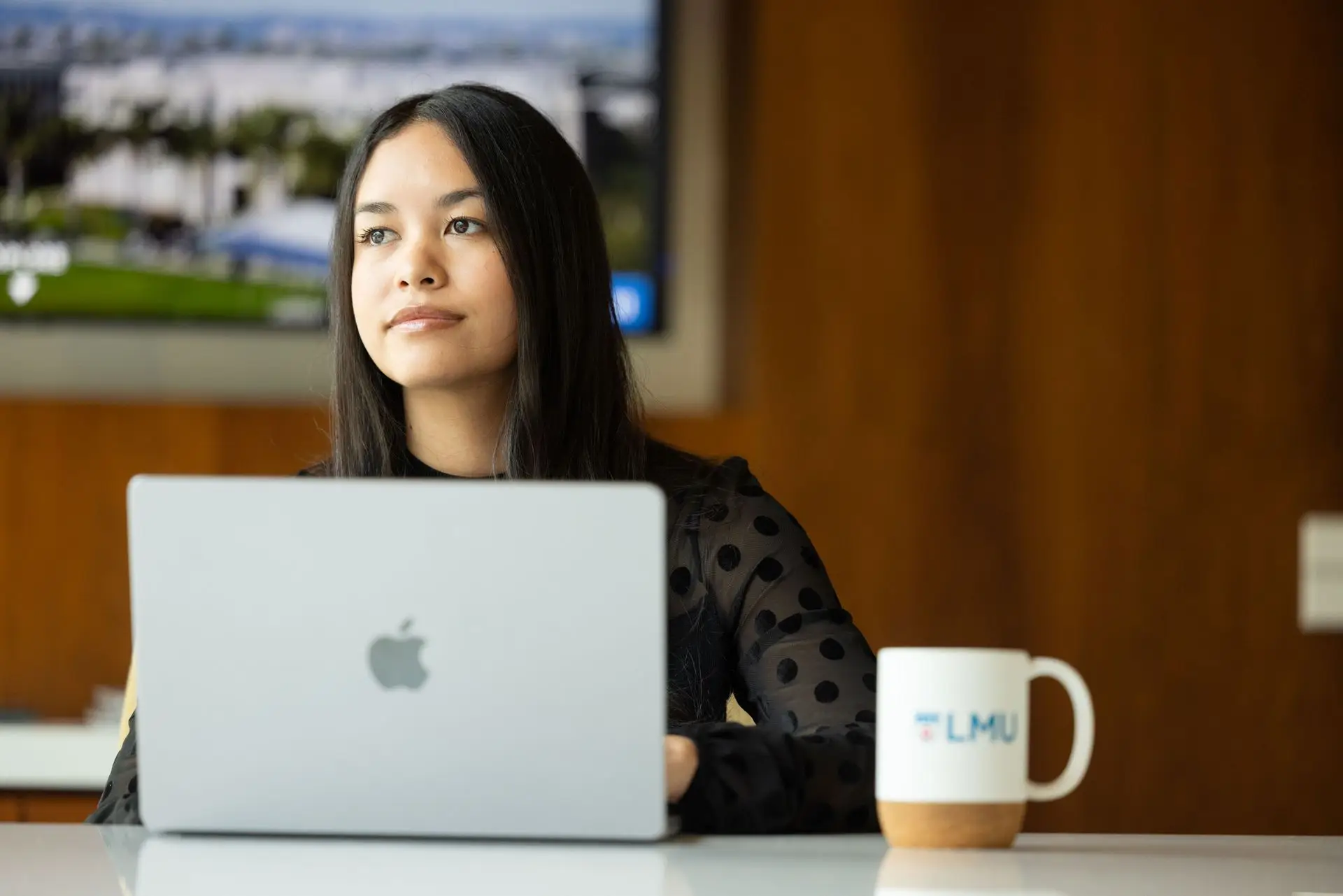 A young woman works on a laptop at a desk, with an LMU mug beside her and campus imagery visible on a screen behind her.